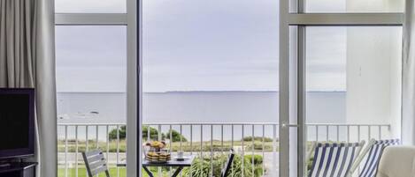 Een balkon met een glazen deur, een televisie, een tafel met stoelen en uitzicht op zee.