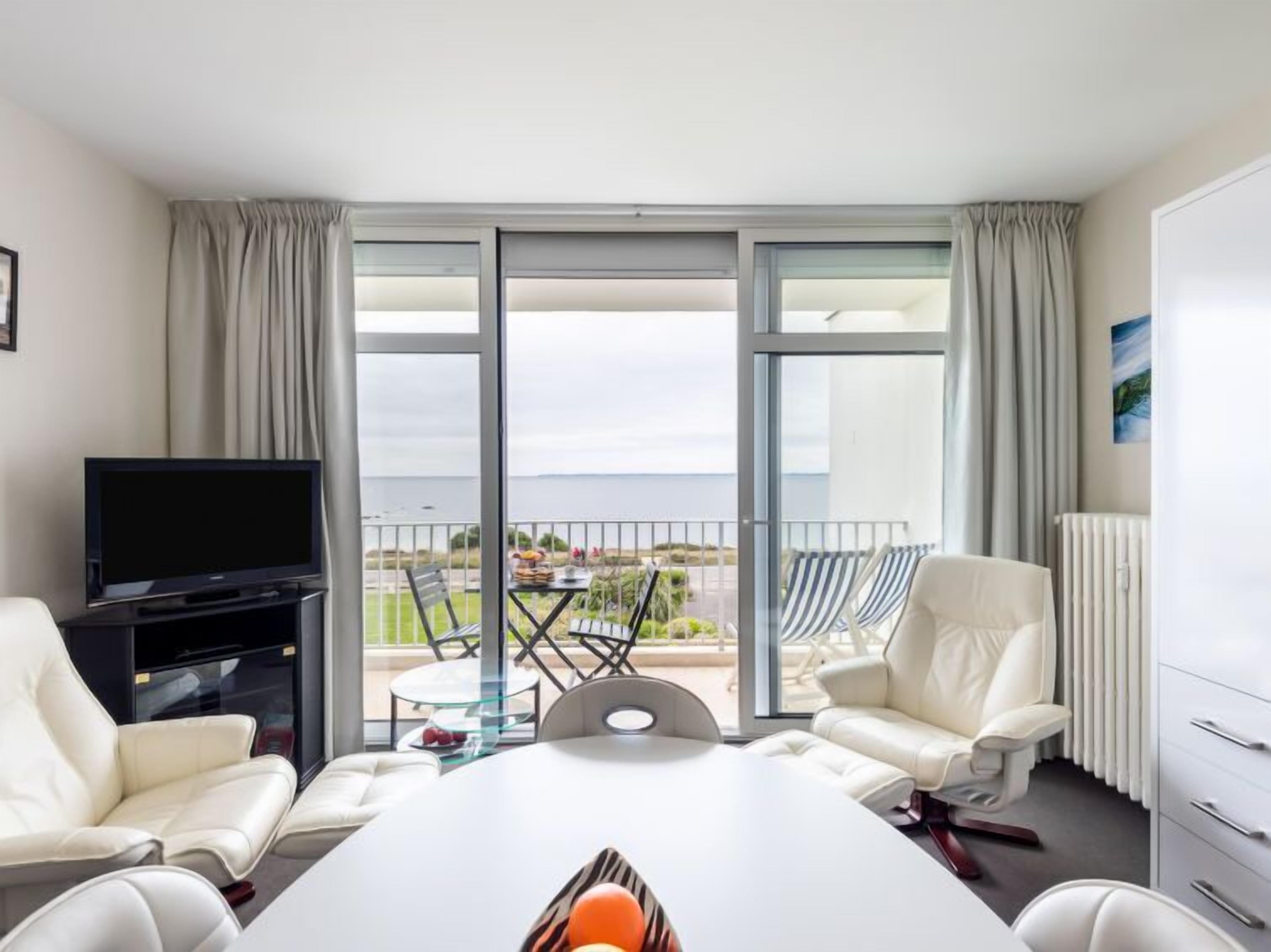 Een hotelkamer met balkon, een televisie, een eettafel en twee witte leren banken.