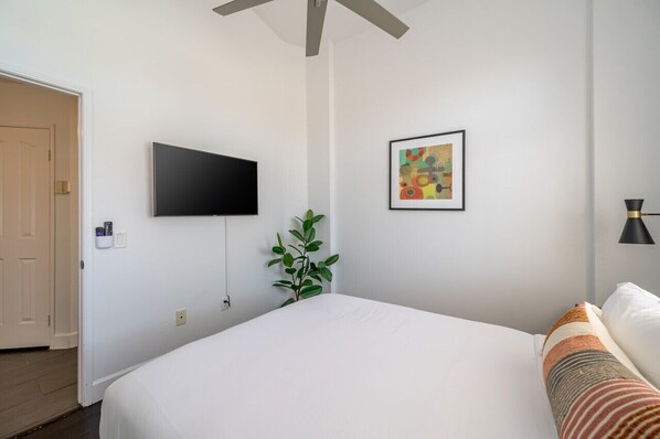 A bedroom with a bed, a television mounted on the wall, a framed picture, a plant, and a door. - Palm Tree Inn | Gated Pool + Large Backyard [Free Spa Heating + Early Check-In] (La Quinta)