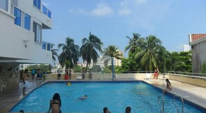 Une piscine o ù des gens nagent et se détendent, entourée de palmiers et de bâtiments.