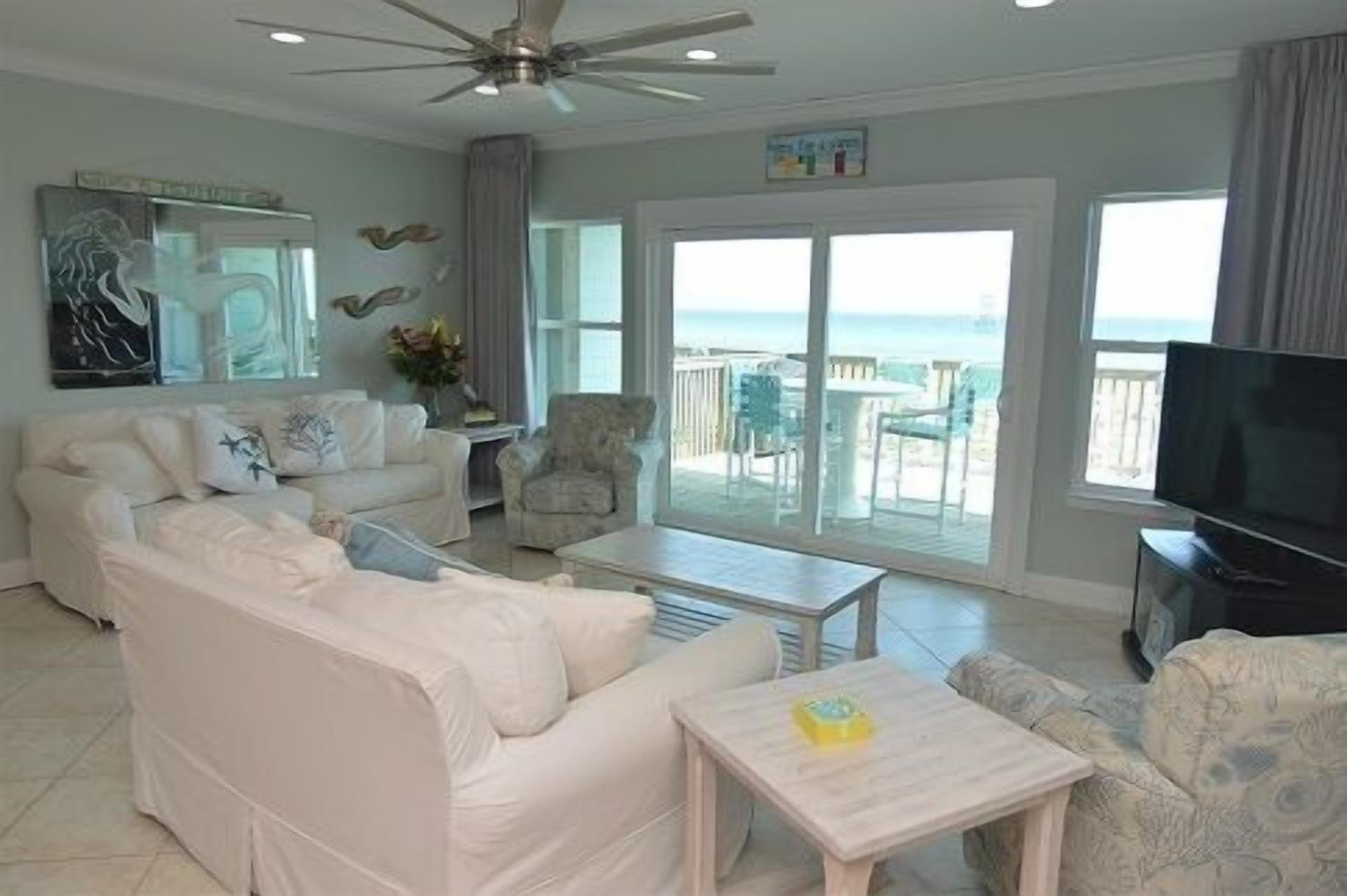 A living room with a white sofa, a coffee table, and a TV. There is a balcony with a view of the ocean.