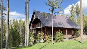 Une cabane en rondins surmontée d’un toit en métal, entourée de grands arbres et d’une pelouse bien entretenue.