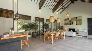 A spacious living area with a dining table, chairs, and a sofa. There are potted plants and a large window providing a view of the outdoors. - Villa Alice Satu by Villa Finder (Seminyak)