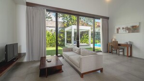 A modern living room with a sofa, a coffee table, a TV, and a dining area with a table and chairs. Large windows offer a view of a garden and a pool. - Villa Alice Satu by Villa Finder (Seminyak)