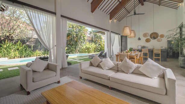 A spacious living area with a large sofa and armchairs, a wooden coffee table, and a view of a swimming pool and lush greenery. - Villa Alice Satu by Villa Finder (Seminyak)