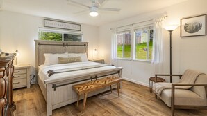Ein Schlafzimmer mit Bett, Nachttischen, einer Bank, einem Sessel, einem Deckenventilator und einem Fenster mit Vorhängen.