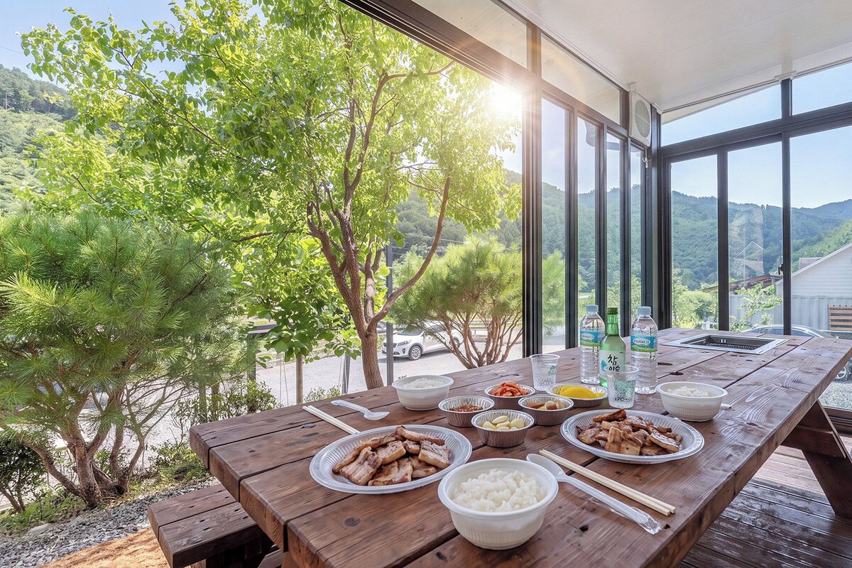 A wooden dining table with various dishes, including rice, meat, and vegetables, set outdoors with a view of a forest and mountains.