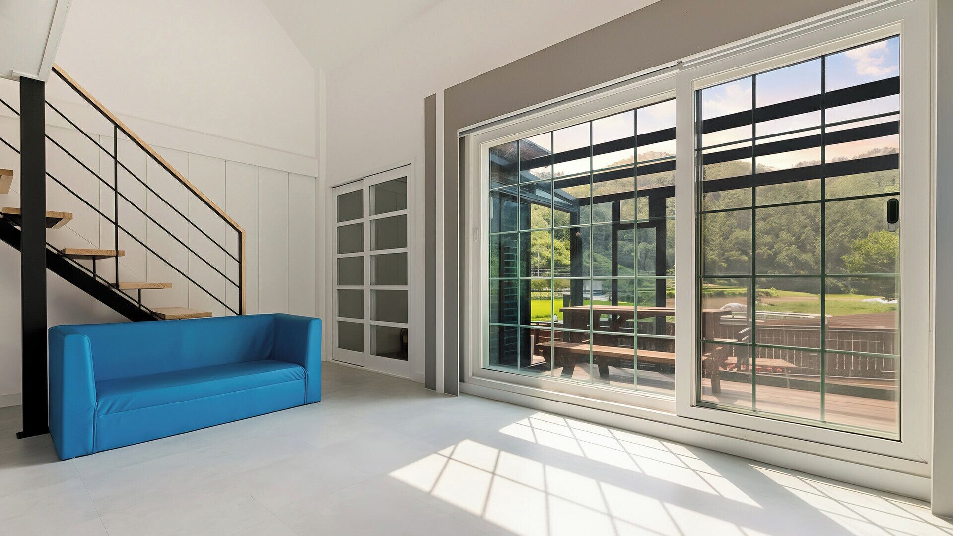 A spacious room with a blue sofa, a staircase with black railings, and large windows offering a view of an outdoor area with a table and chairs.