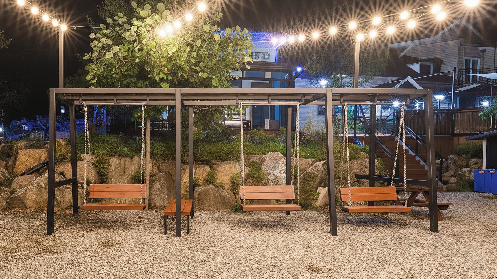 Une balançoire en bois avec des bancs, entourée de rochers et de gravier, illuminée par des guirlandes lumineuses la nuit.