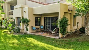 A covered patio with outdoor furniture, a small garden, and a building with a beige facade.