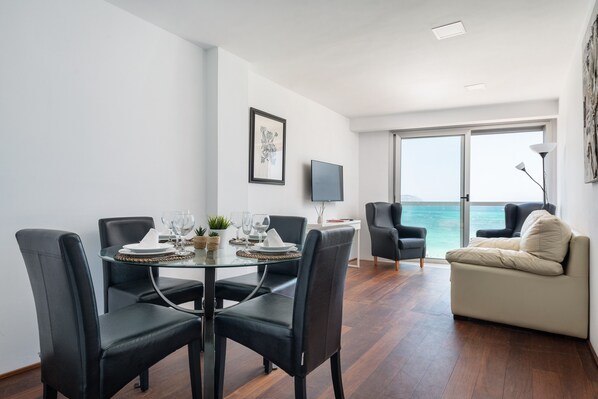 A dining area with a glass table, black chairs, and a sofa facing a large window with a view of the sea.
