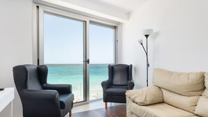 Een kamer met een grote schuifglazen deur die uitkomt op een uitzicht op het strand, twee zwarte leren fauteuils, een beige bank en een vloerlamp.