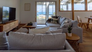 Un salon moderne avec un canapé gris, un téléviseur à écran plat posé sur un meuble en bois, et une vue sur la mer grâce à de grandes portes coulissantes.