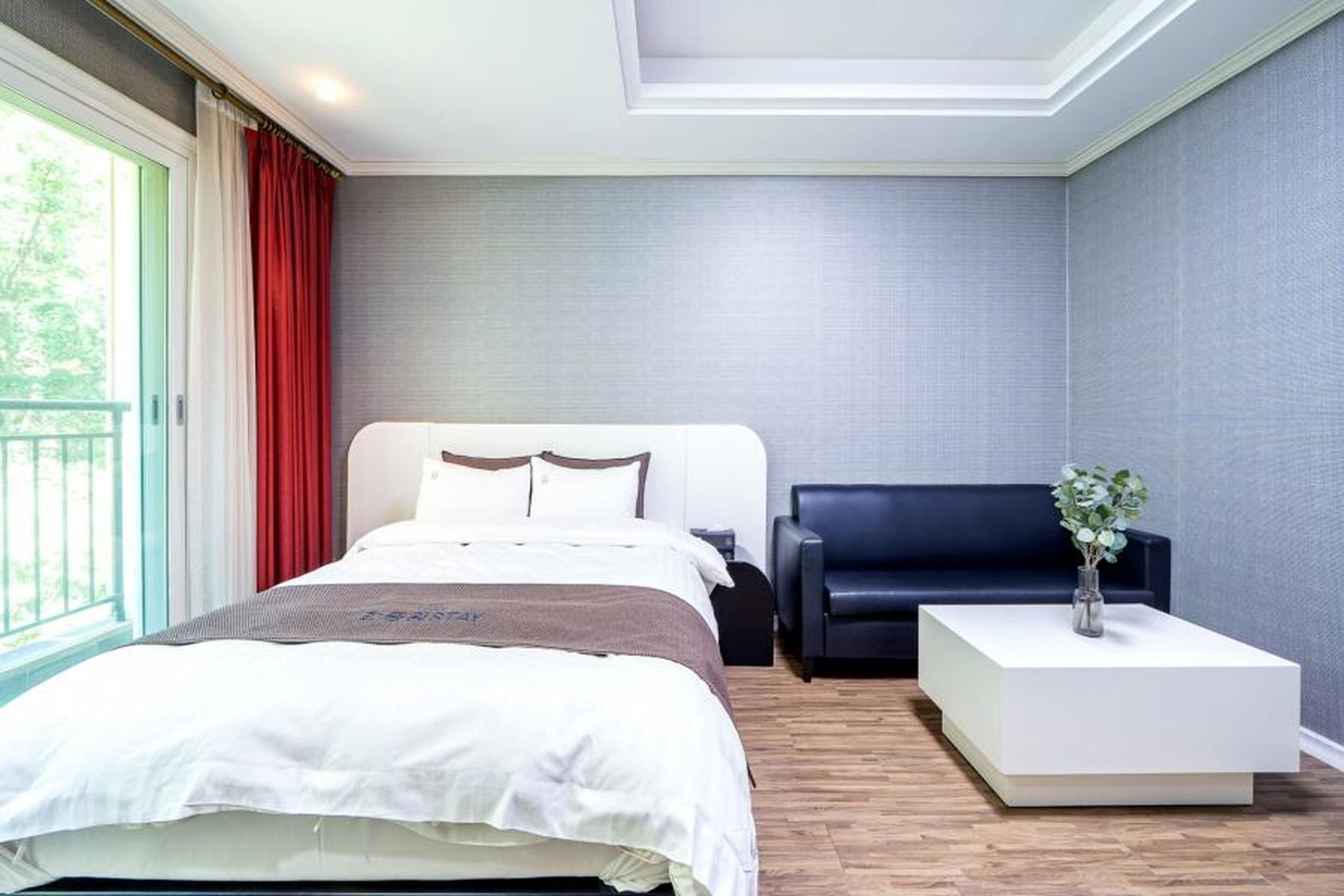Una habitación de hotel moderna con una cama grande, un sofá oscuro, una mesita de noche blanca y vistas a la vegetación por la ventana.