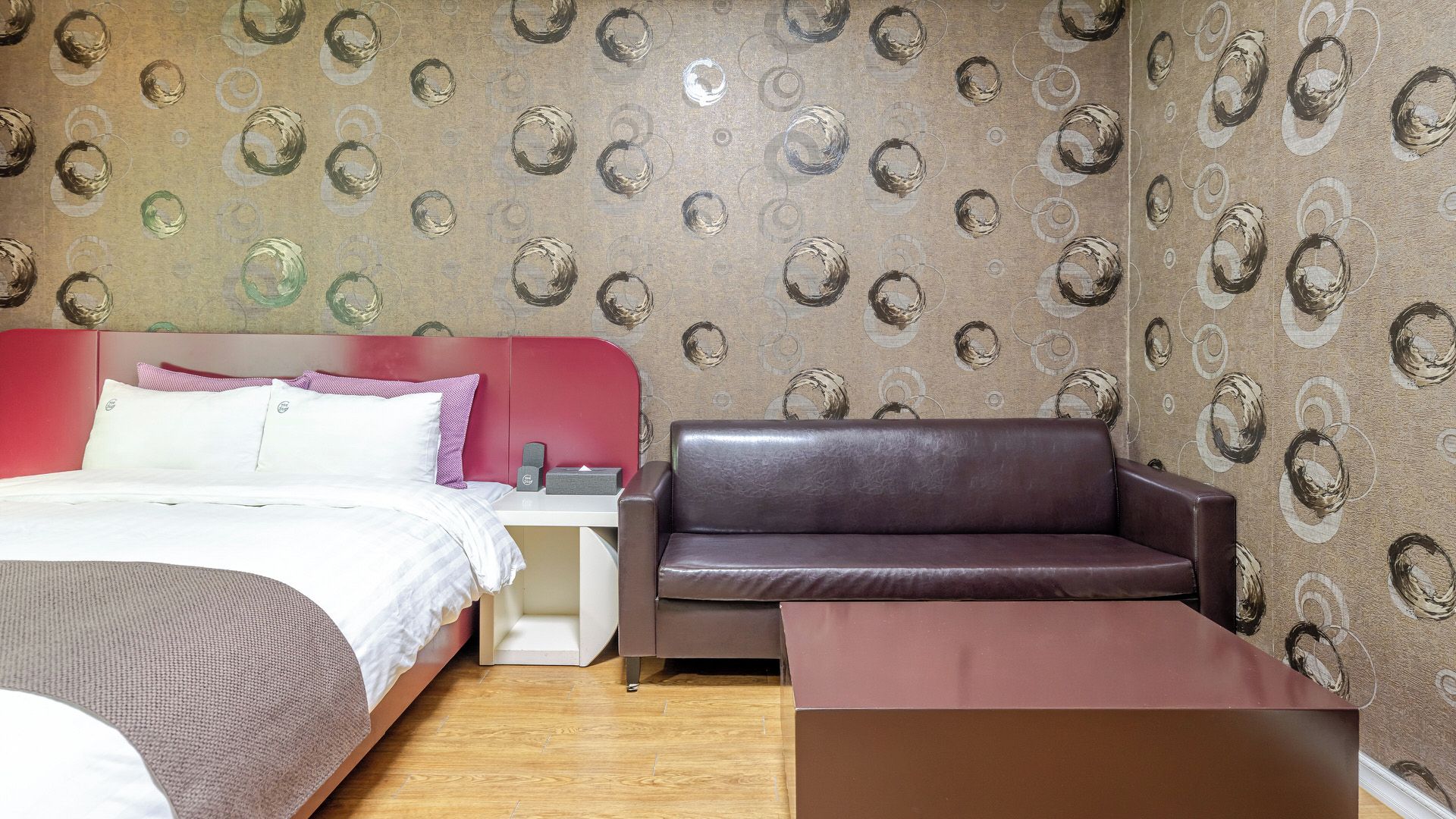 A hotel room with a bed, a red headboard, a brown leather sofa, a white nightstand, and a patterned wallpaper.