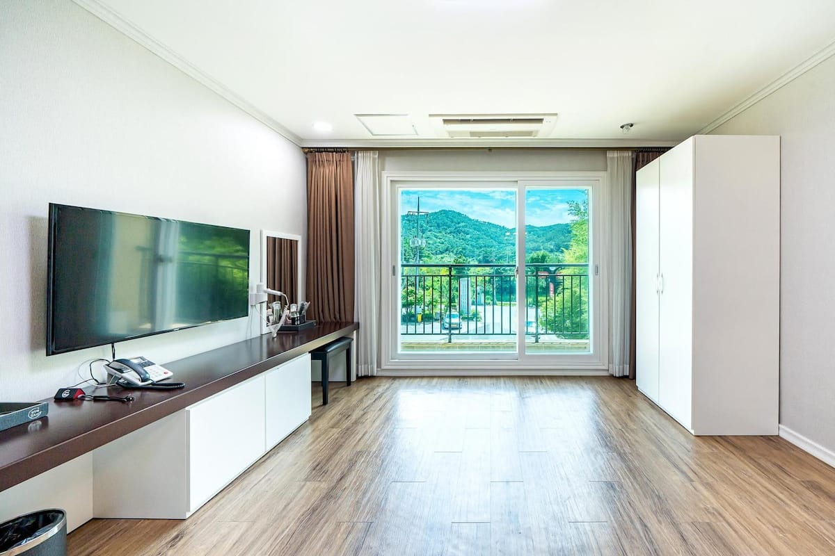 Ein modernes Hotelzimmer mit einem Flachbildfernseher, einem Schreibtisch mit Telefon und Blick auf eine grüne Landschaft durch eine große Schiebeglastür.