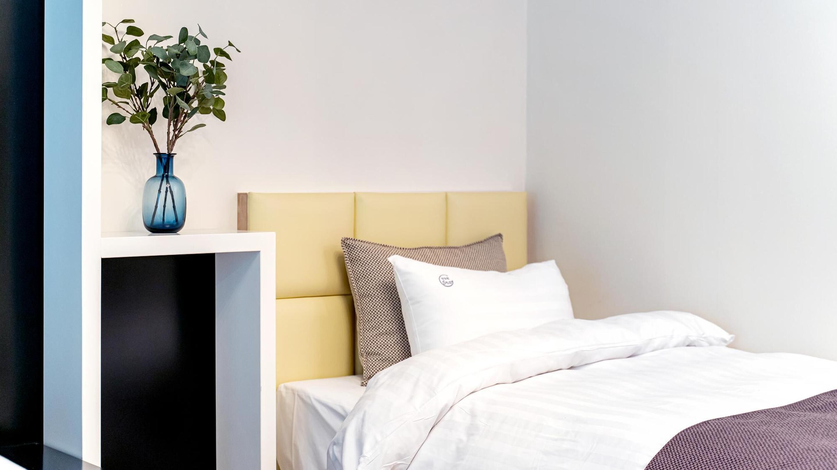Habitación de hotel con cama, mesita de noche, un florero con plantas verdes y cabecero con almohadas.