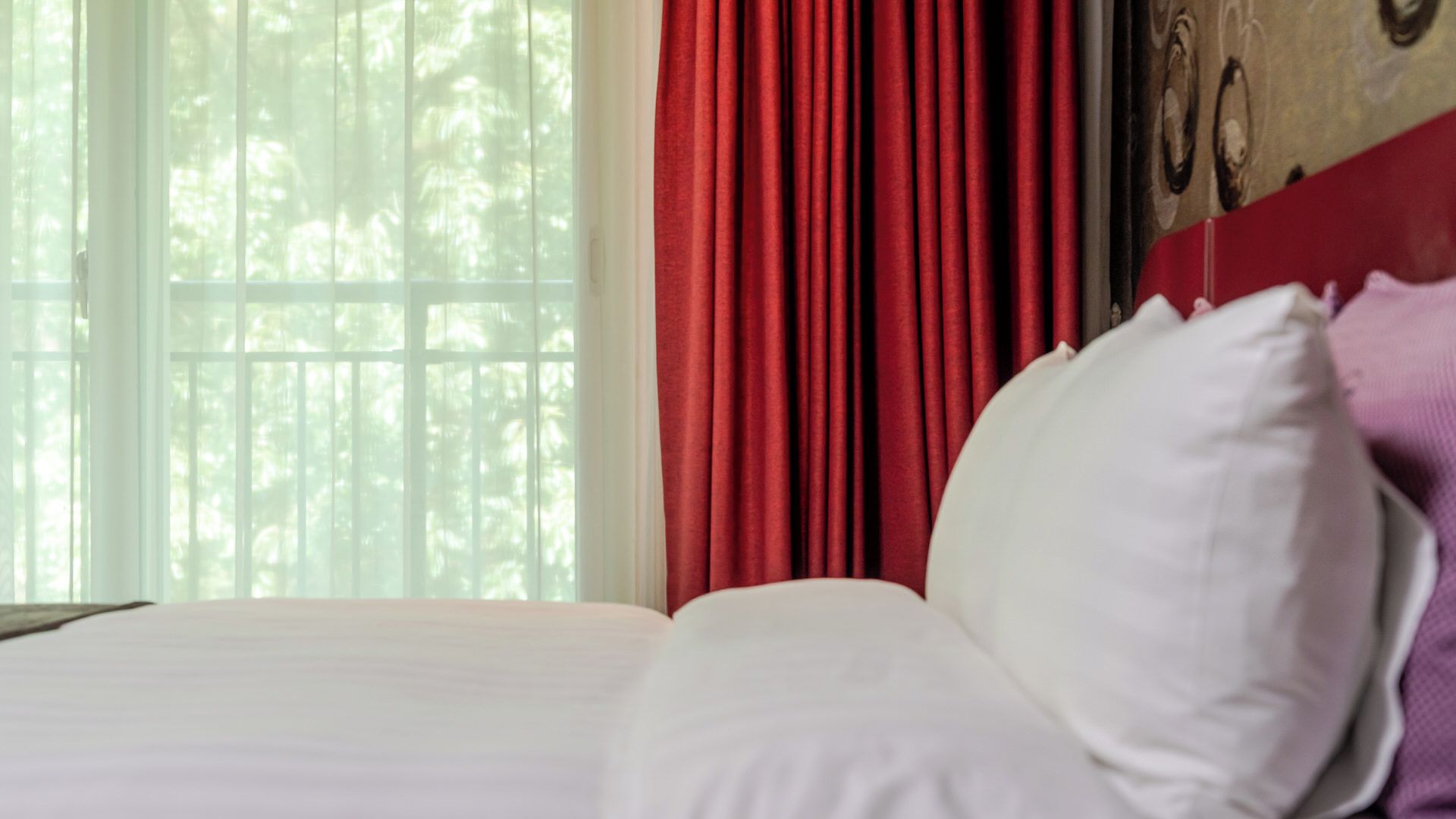 A bed with white bedding, a red curtain, and a window with a view of trees.