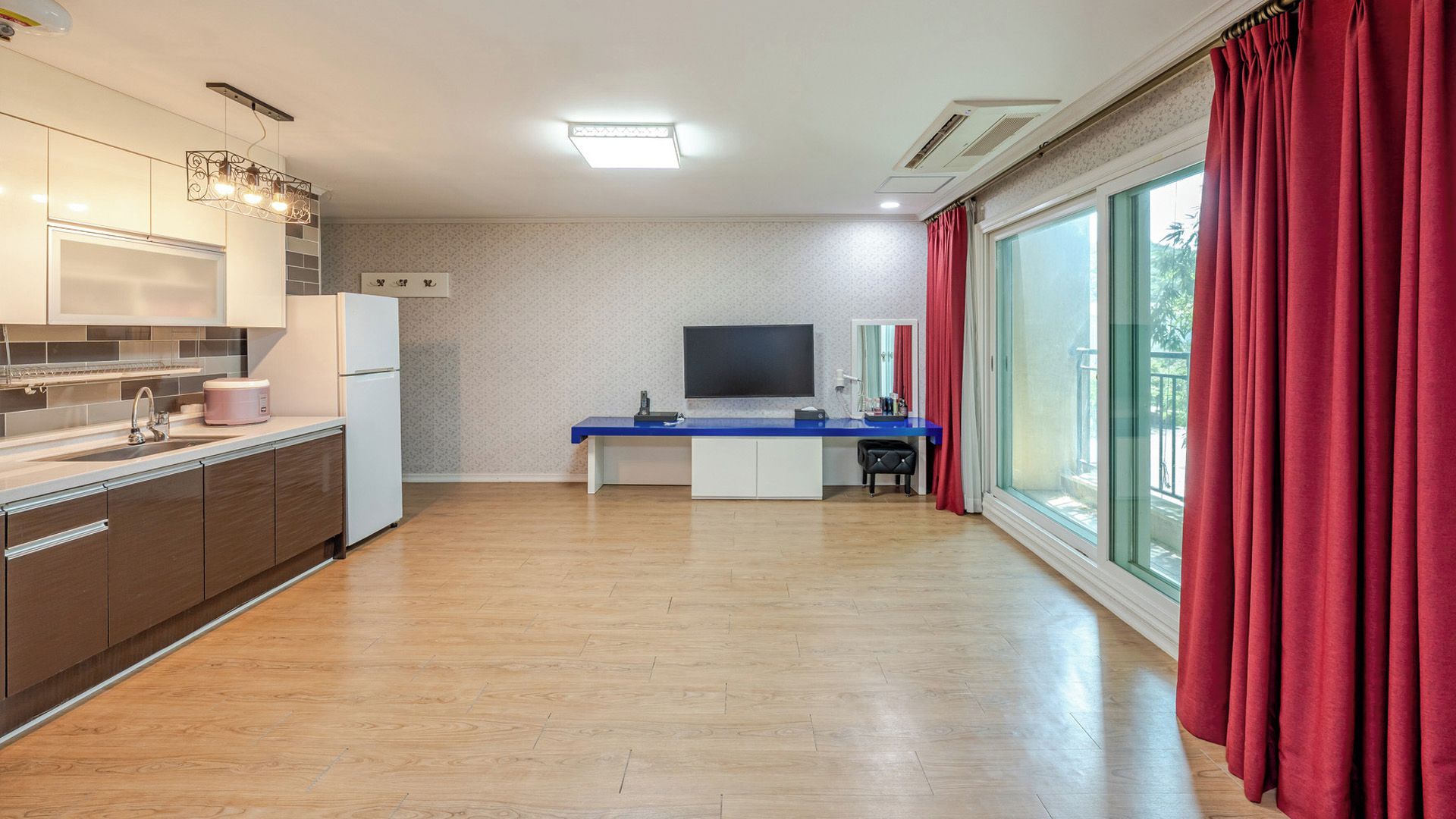 A modern kitchen with a built-in oven, a dining area with a blue table, and a large window with red curtains.