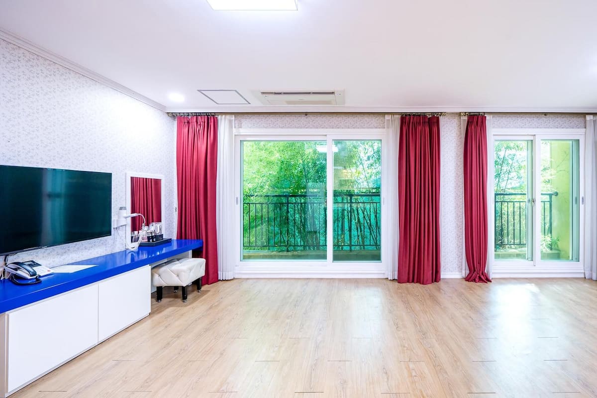 Ein geräumiges Zimmer mit einem großen Schiebeglasfenster, das auf einen Balkon führt, einer blauen Ablage mit Telefon und Kaffeemaschine sowie einem an der Wand befestigten Flachbildfernseher.