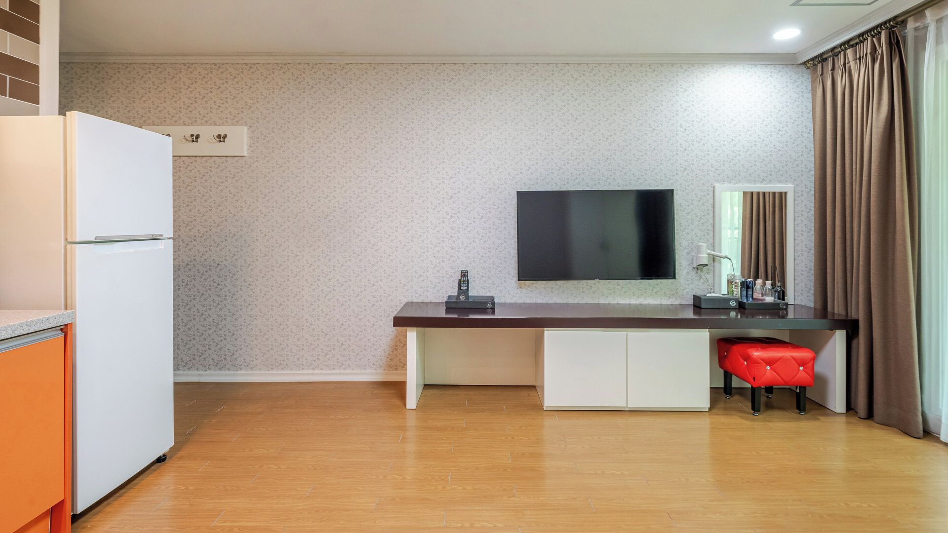 Una cocina compacta con refrigerador, un televisor de pantalla plana montado en la pared y una mesa de comedor de madera con una silla roja.