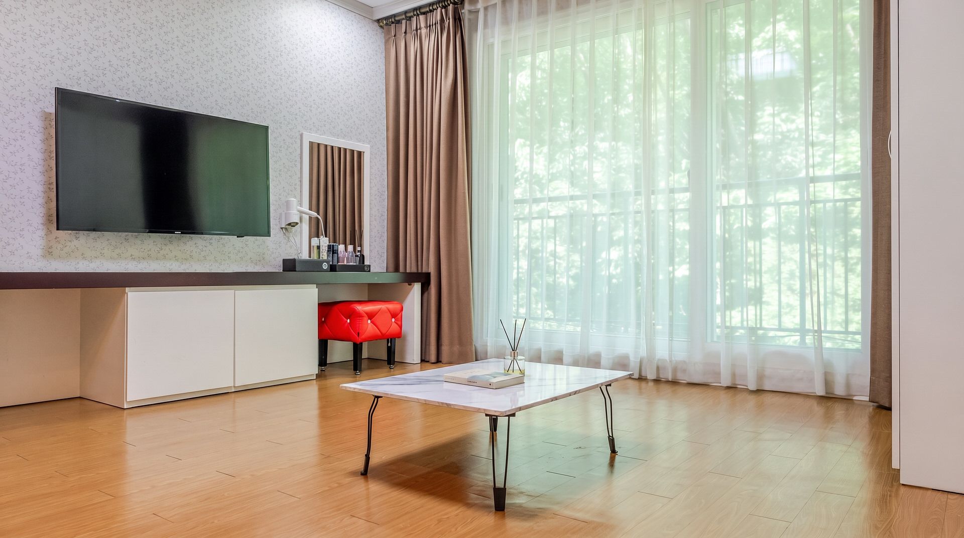 A modern living room with a flat-screen TV, a white coffee table, a red chair, and a large window with sheer curtains.