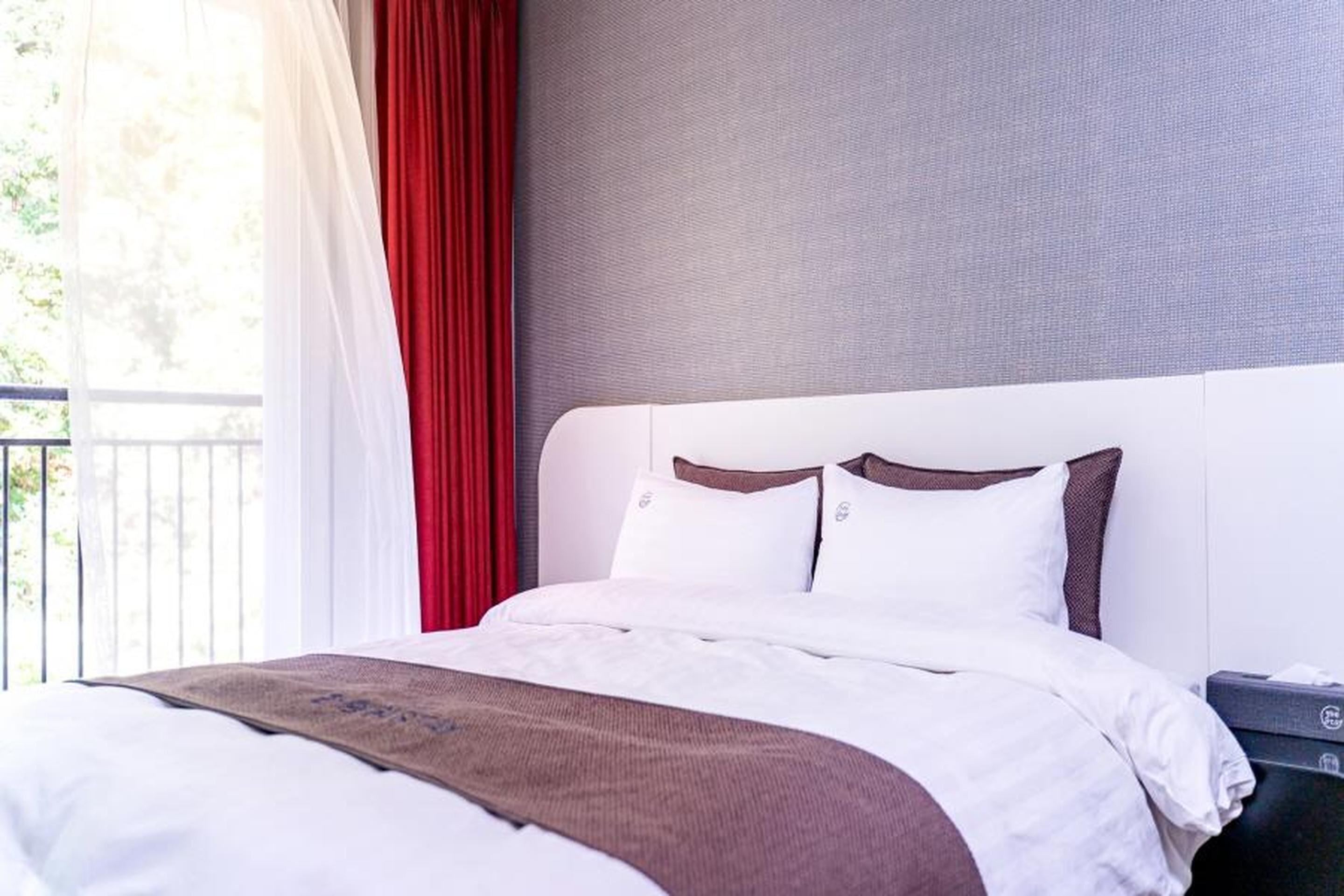 A hotel room with a bed, a nightstand, and a window with red curtains.