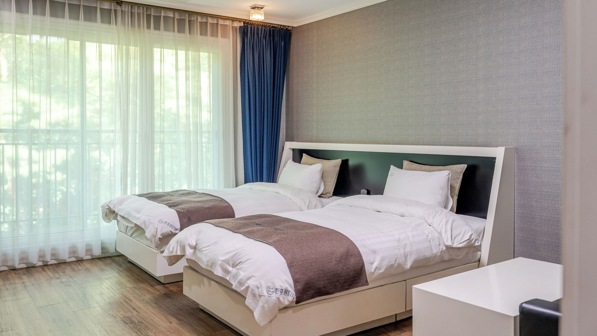 A double bed with a headboard, a bedside table, and a window with sheer curtains.