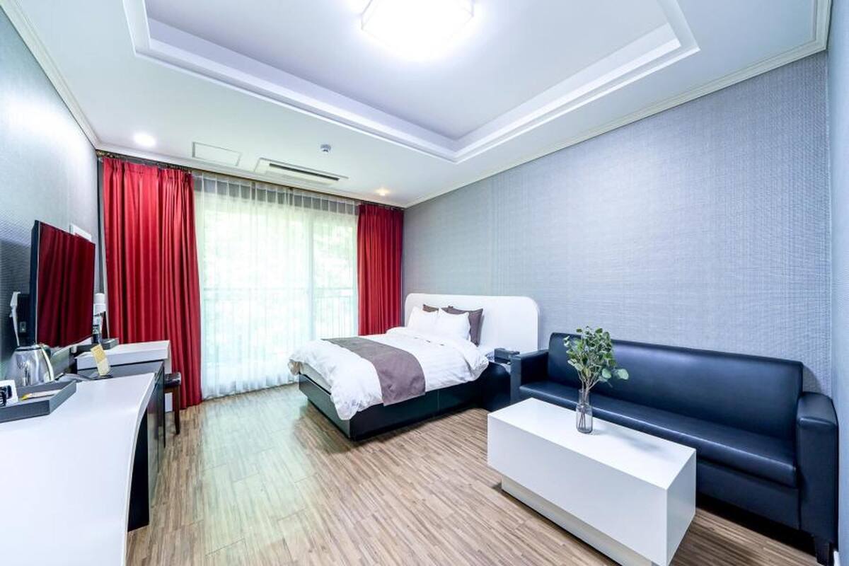 Ein modernes Hotelzimmer mit Bett, Schreibtisch, Sofa und einem großen Fenster mit roten Vorhängen.