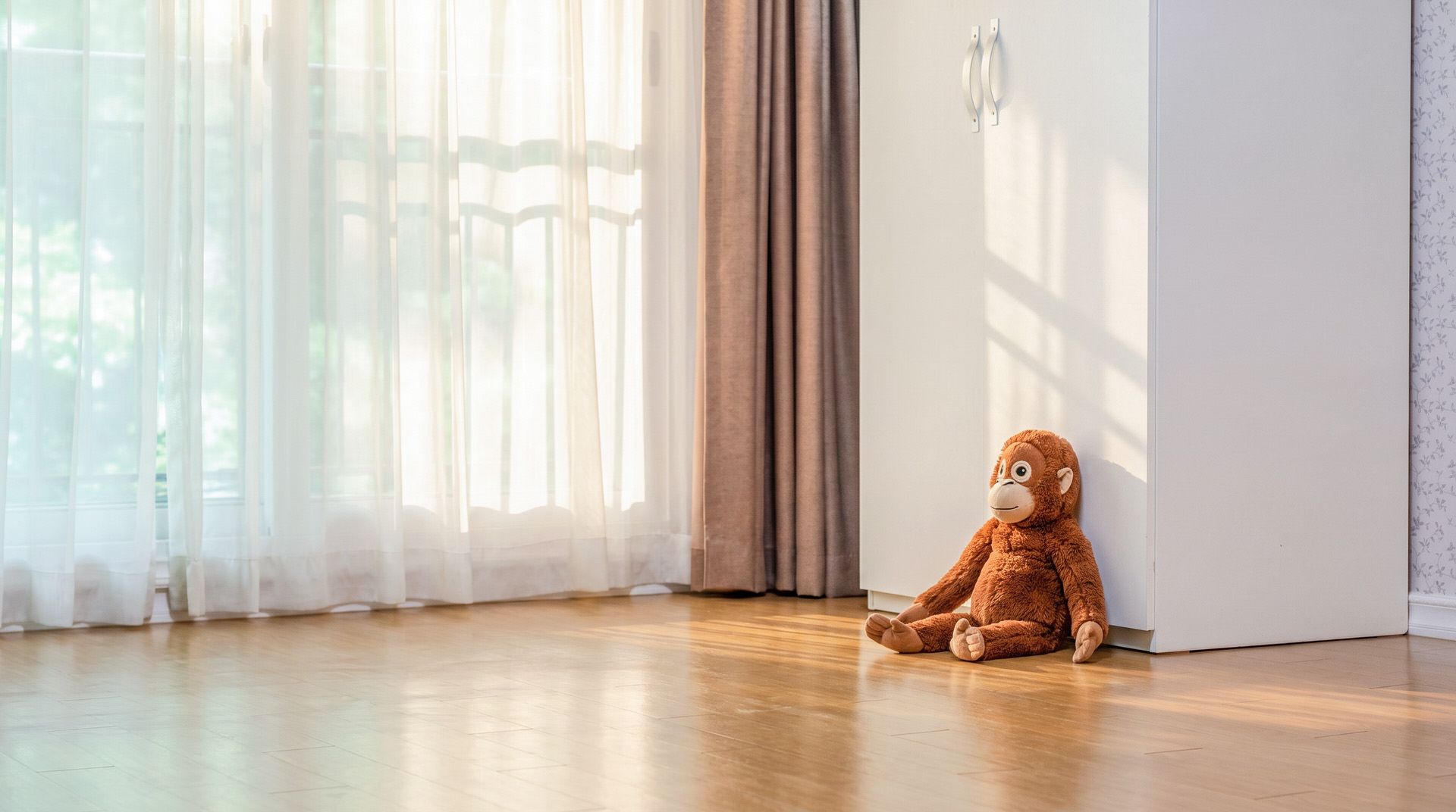 A room with a wooden floor, a large window with sheer curtains, and a plush toy sitting by the door.