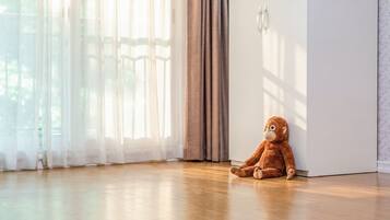 A room with a wooden floor, a large window with sheer curtains, and a plush toy sitting by the door.