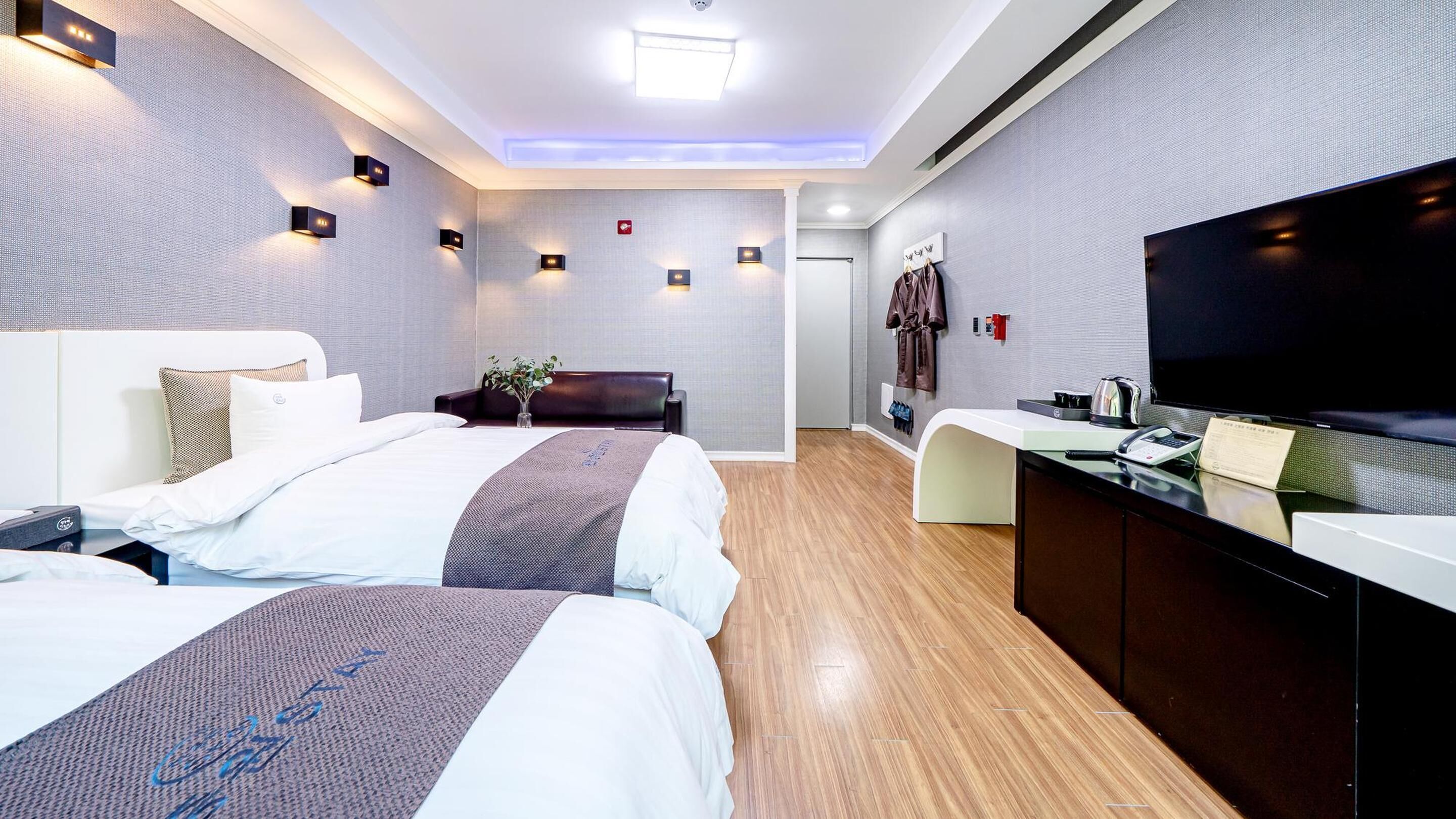 A modern hotel room with two beds, a desk, a flat-screen TV, and a reception area.