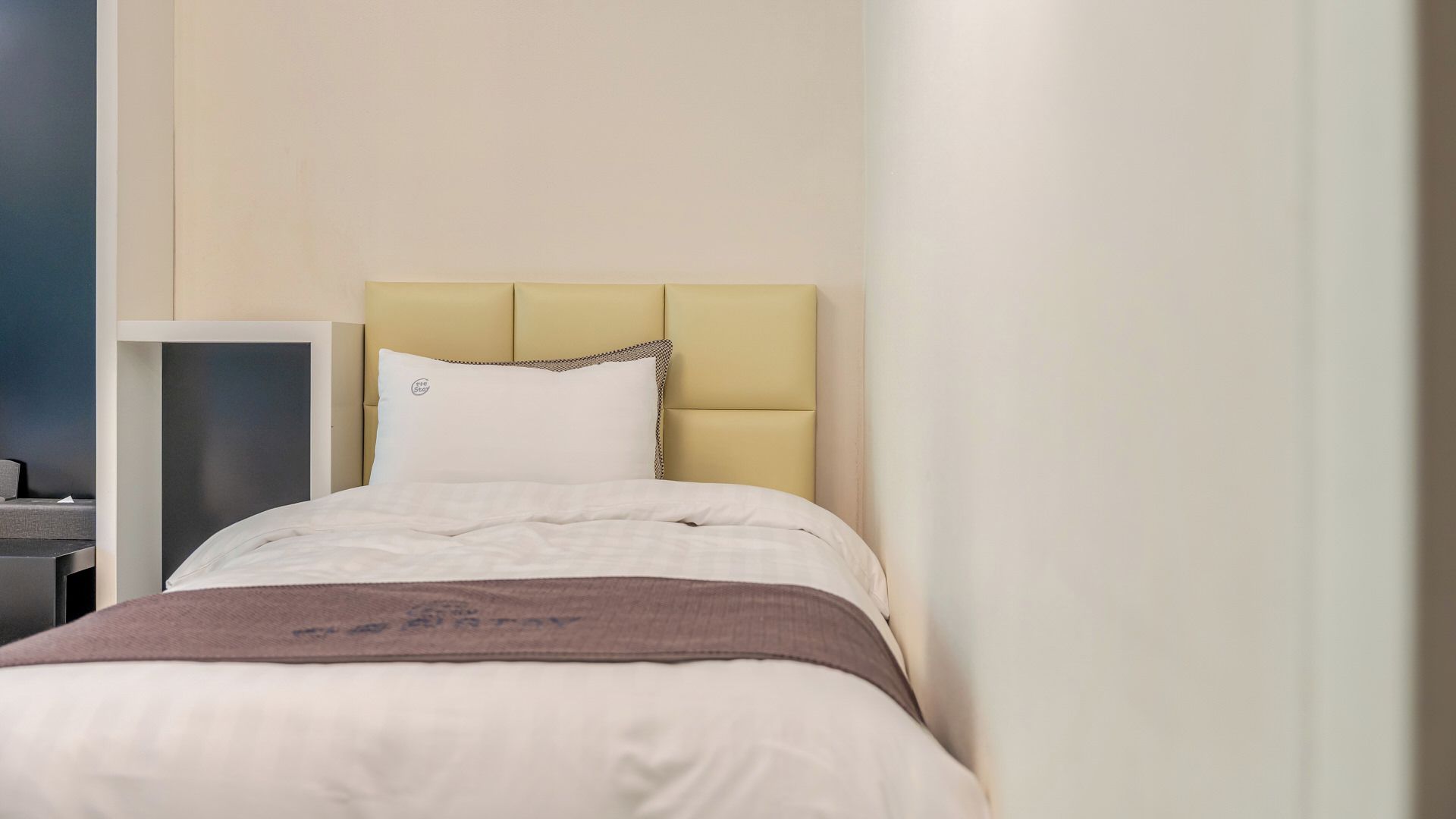 Una cama bien hecha con cabecera, mesita de noche y estante en la pared.
