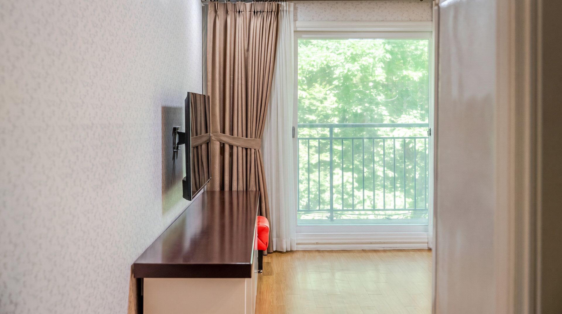 A room with a TV mounted on the wall, a window with curtains, and a balcony with a railing.