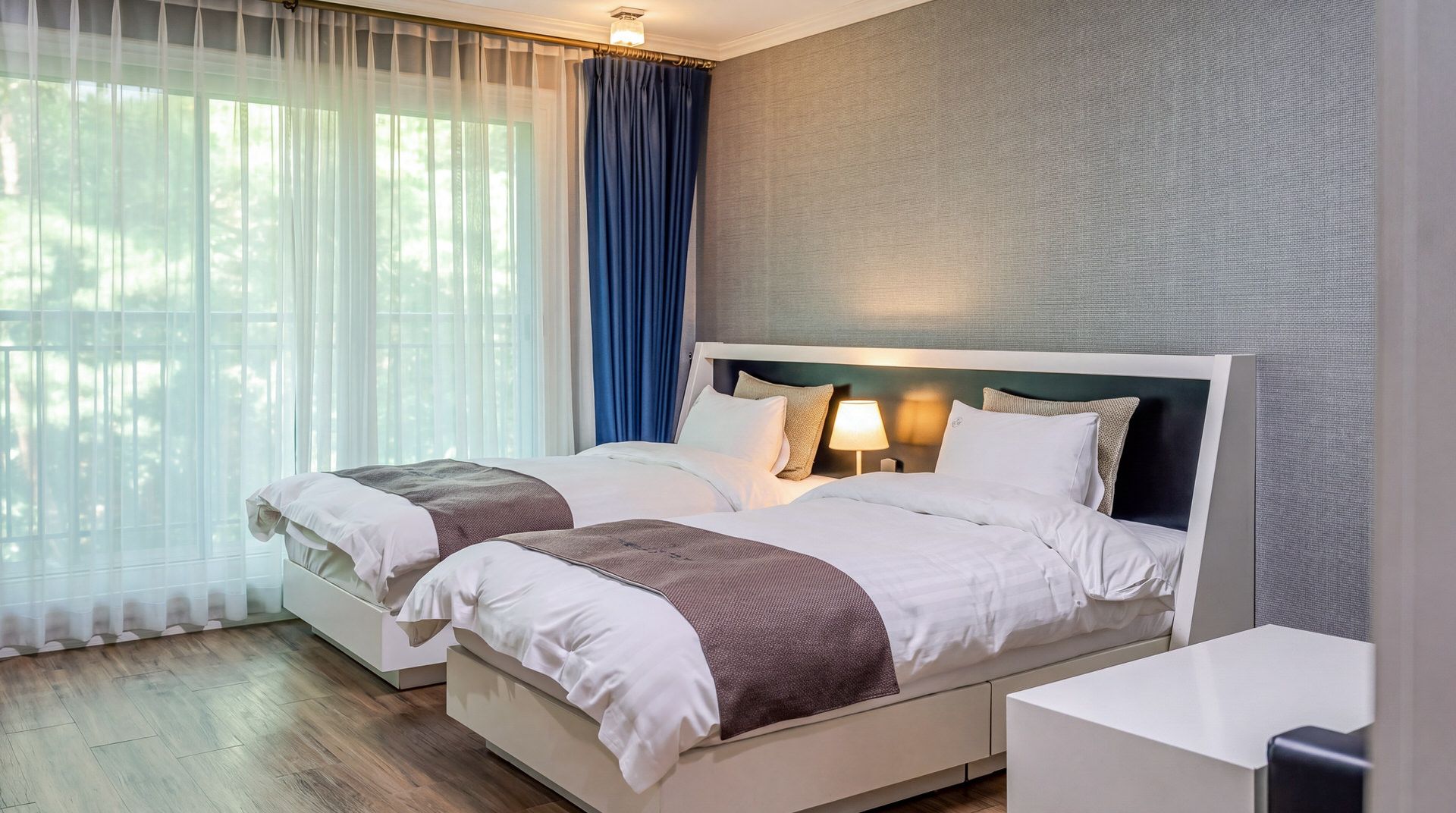 A double bed with a headboard, two bedside tables, a lamp, and a view of greenery through sheer curtains.