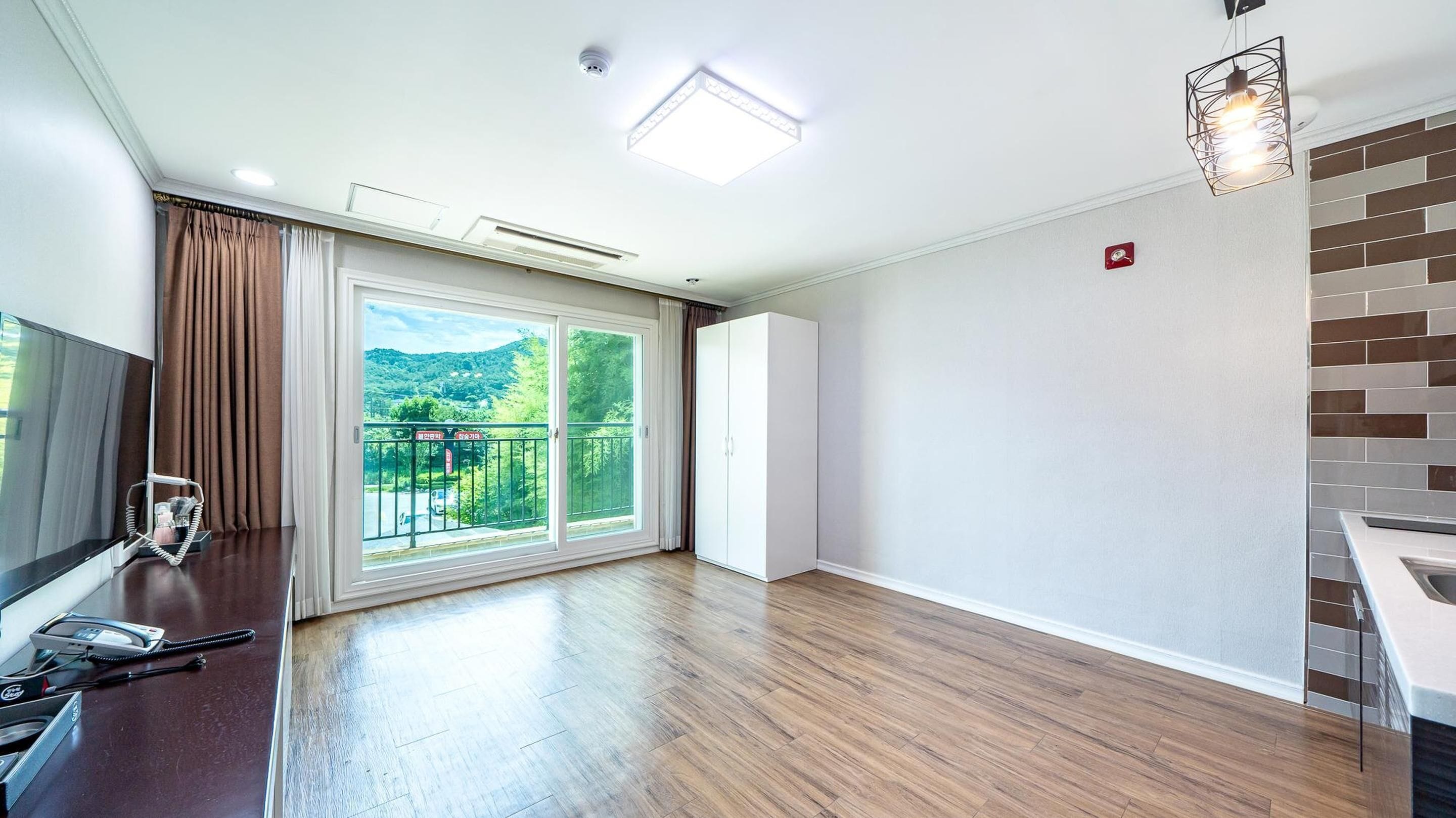 Una habitación amplia con un gran ventanal corredero que da a un balcón, suelo de madera, un armario blanco y una lámpara moderna.