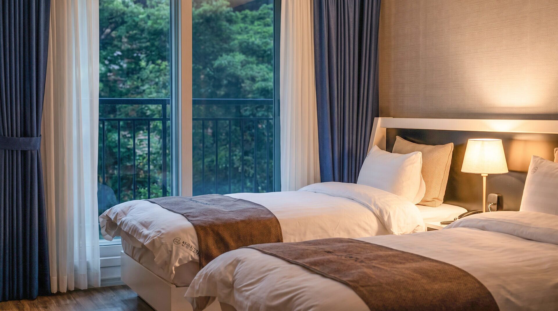 A hotel room with two beds, a balcony, and a view of greenery.