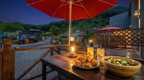 An outdoor dining area with a wooden table, a red umbrella, a lantern, and a bowl of salad.