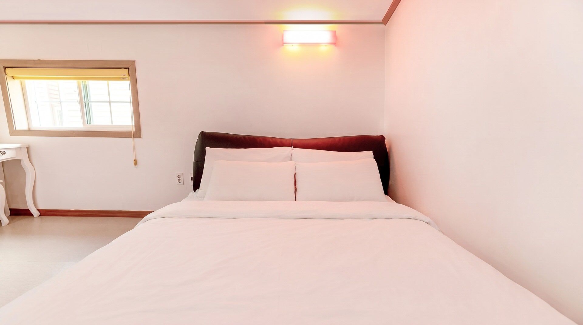 A bedroom with a bed, a nightstand, a window with blinds, and a wall-mounted light fixture.