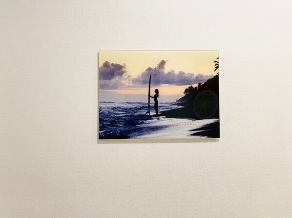 Ein gerahmtes Bild zeigt eine Strandansicht mit einer Person, die ein Surfbrett hält, einer großen Limette und einem Sonnenuntergang im Hintergrund.