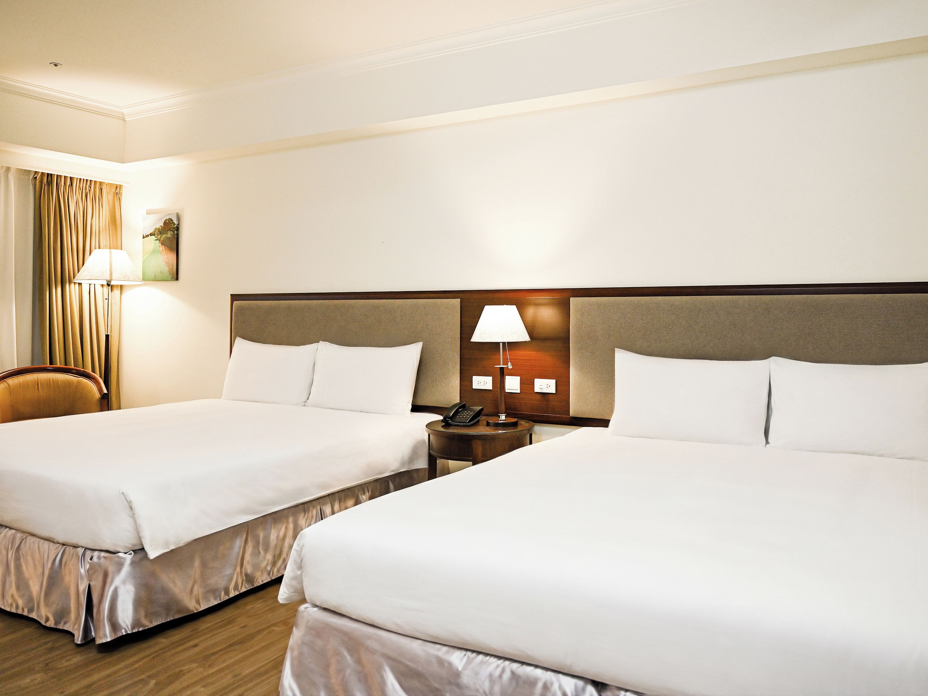 A hotel room with two beds, a nightstand with a lamp, a chair, and a painting on the wall.