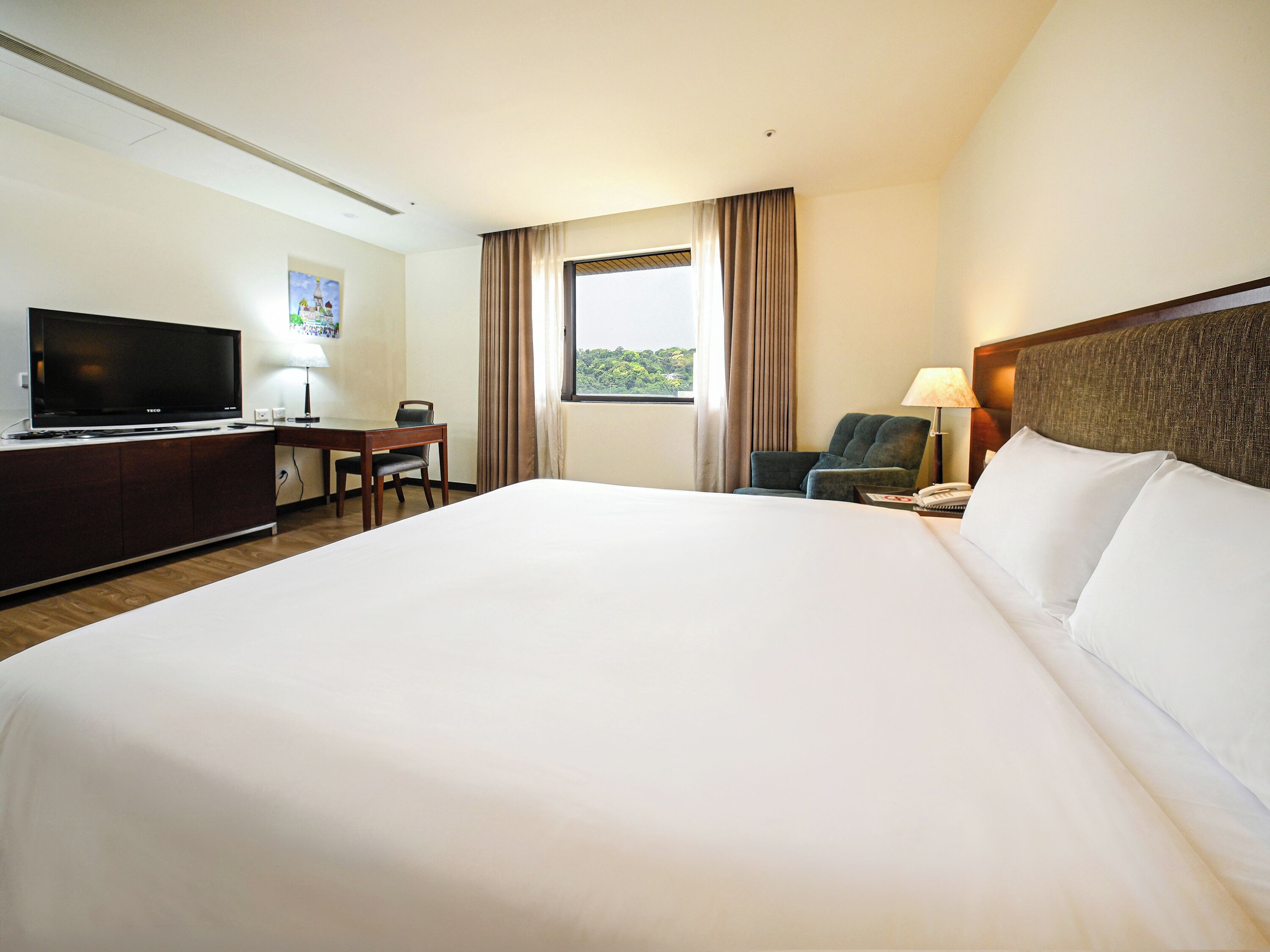 A hotel room with a large bed, a desk with a flat-screen TV, a chair, a window with curtains, and a lamp.
