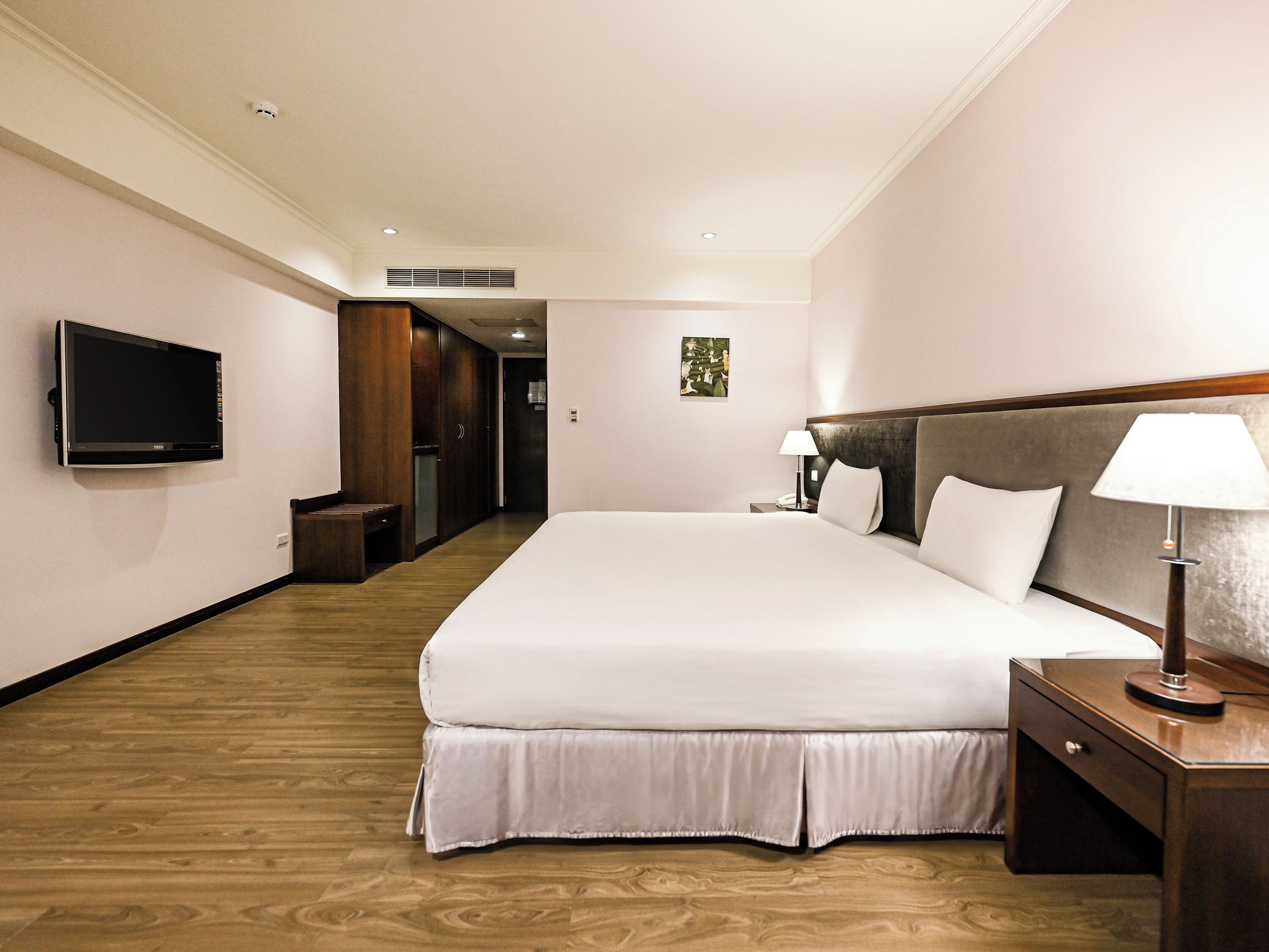 A hotel room with a large bed, a flat-screen TV, a bedside table with a lamp, and a wooden floor.