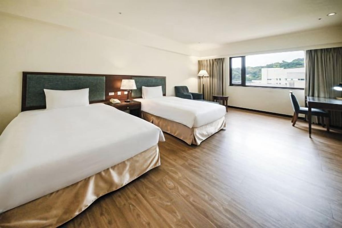Quarto de hotel com duas camas, uma mesa, uma cadeira e janela com vista.