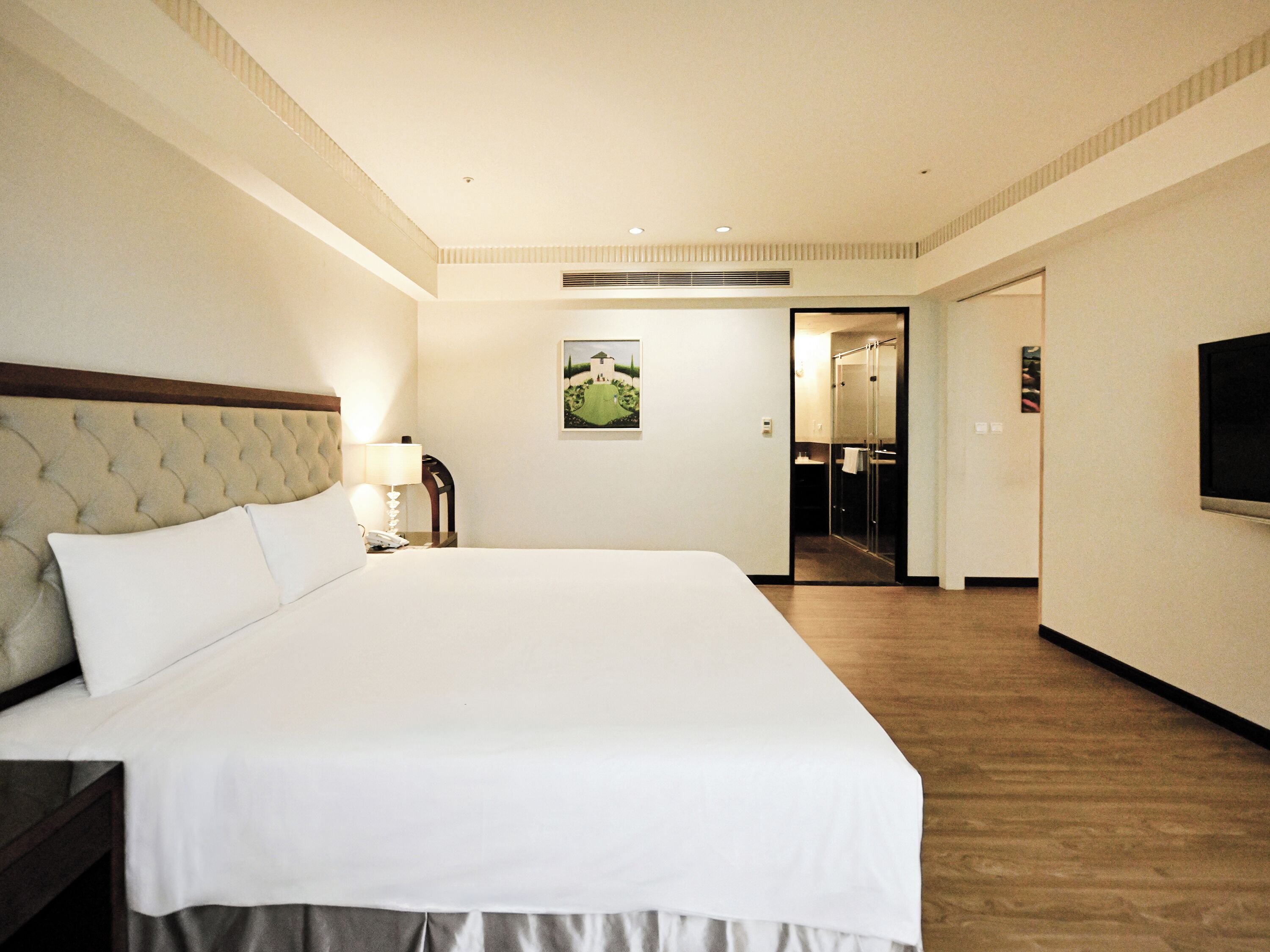Una habitación de hotel con una cama grande, una mesita de noche con lámpara, un cuadro en la pared y un televisor montado en la pared.