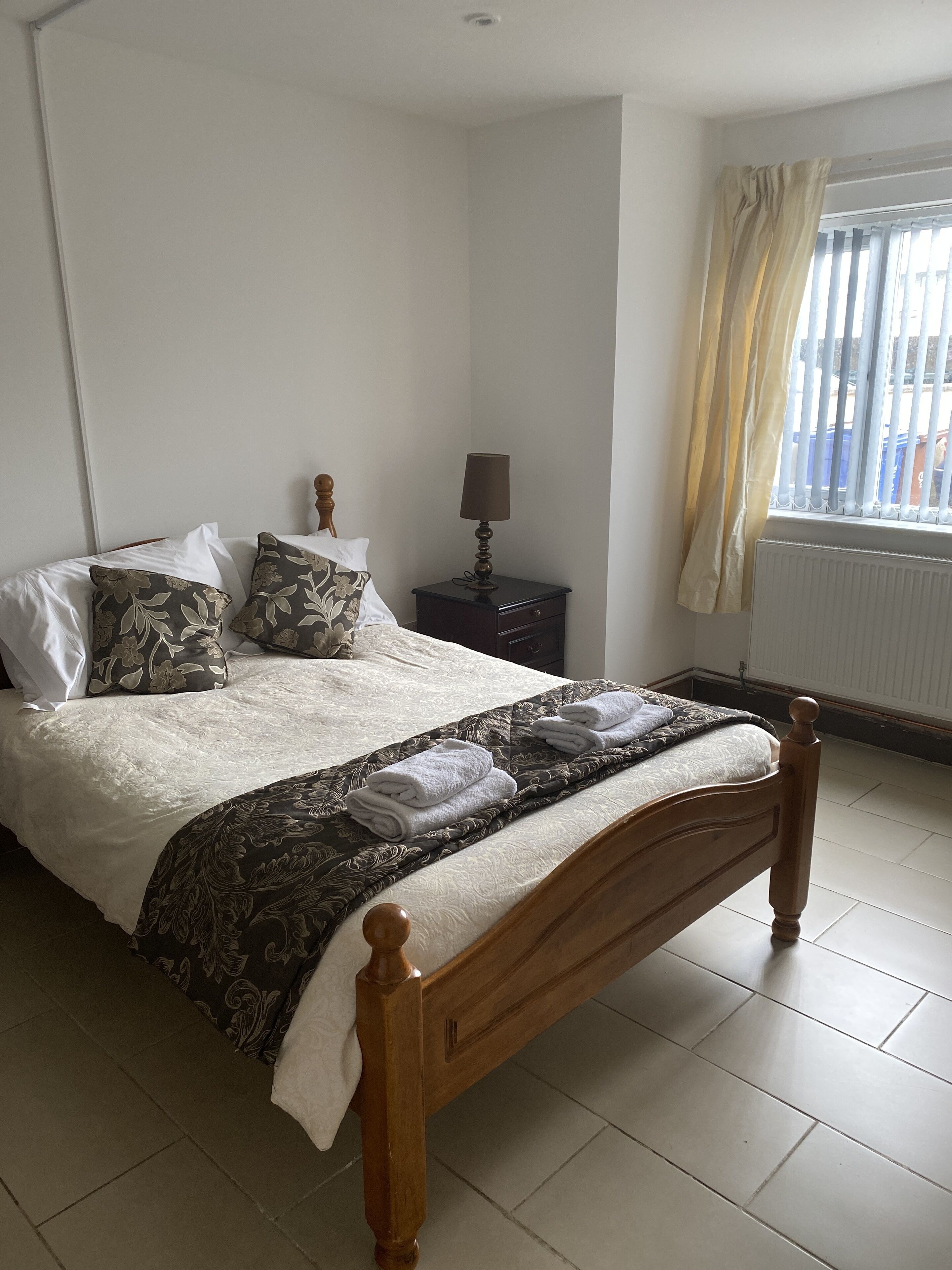 A neatly made bed with a patterned bedspread and pillows, a bedside table with a lamp, and a window with curtains.