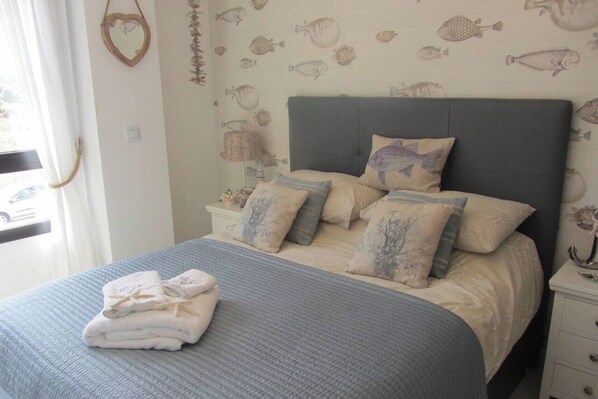 A neatly made bed with a blue bedspread and multiple pillows, a bedside table with a lamp, and a wall with fish patterns. - City Apt - Sleeps 4, Community Pool & Parking (Denia)