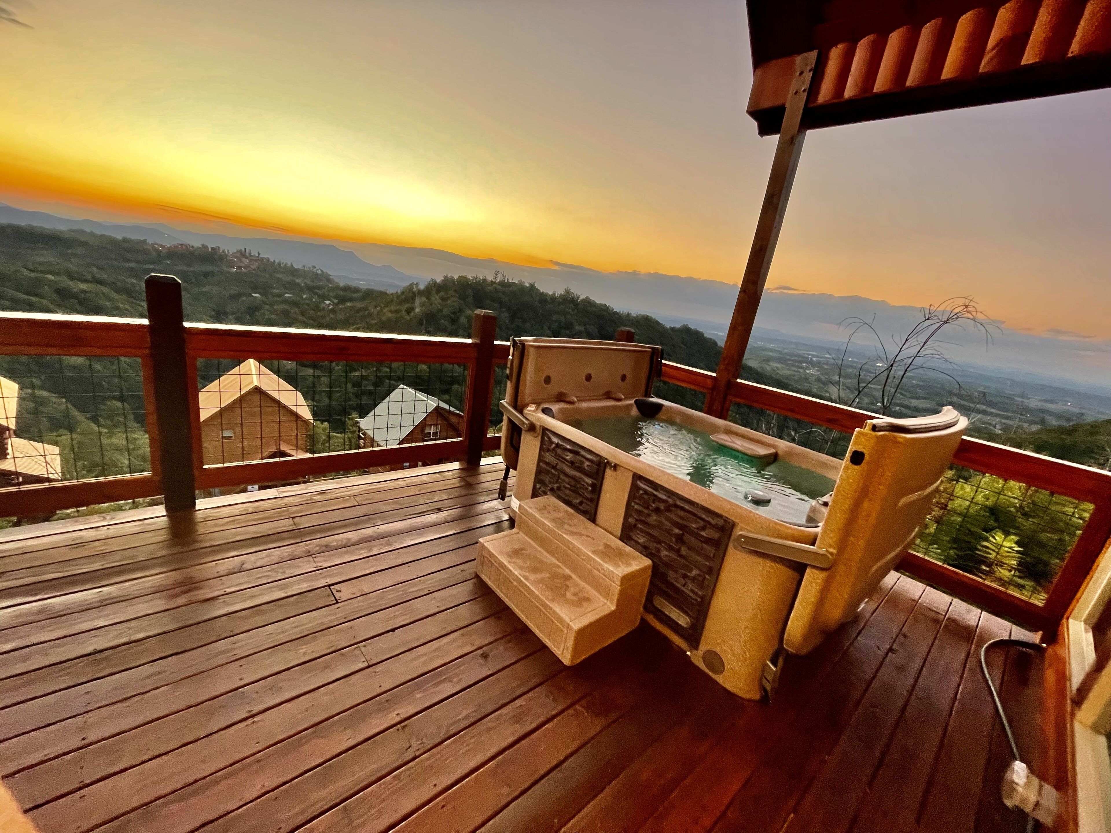 A wooden deck with a hot tub and seating area, overlooking a mountainous landscape during sunset.
