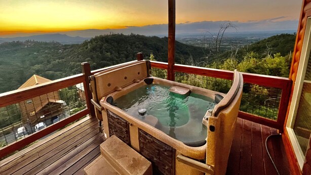 A hot tub on a wooden deck with a view of a forested mountain range during sunset. - Cozy 2bedroom cabin near Island / Dollywood at Pigeon Forge  with Panormic Views (Sevierville)