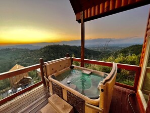 Een hottub op een houten terras met uitzicht op een bebost berglandschap tijdens de zonsondergang.
